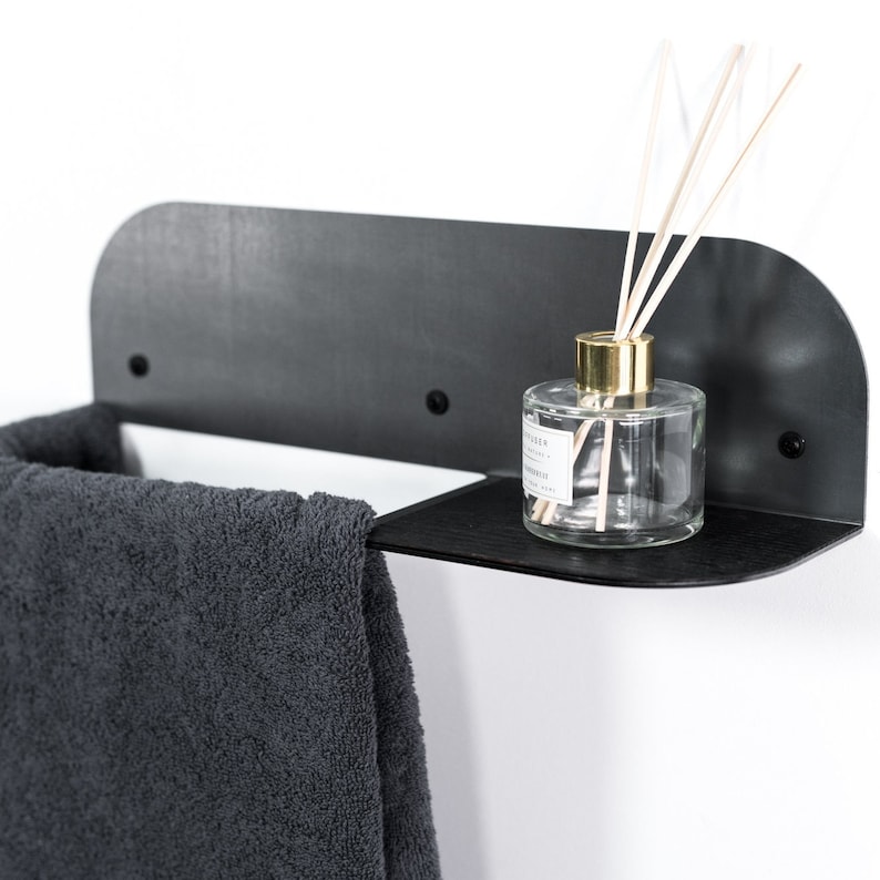 May include: A black metal shelf with a curved edge, mounted on a white wall. A gray towel hangs on the left side of the shelf, and a clear glass bottle with a gold lid and wooden sticks sits on the right side of the shelf.