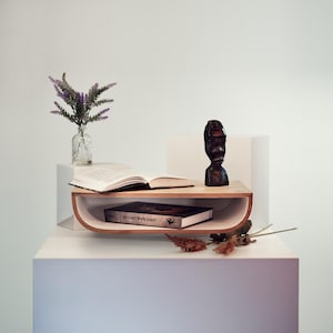 May include: A wooden shelf with a curved front, holding an open book, a closed book, and a small wooden sculpture. A small glass vase with purple flowers sits on a white block. Dried flowers are scattered around the shelf.