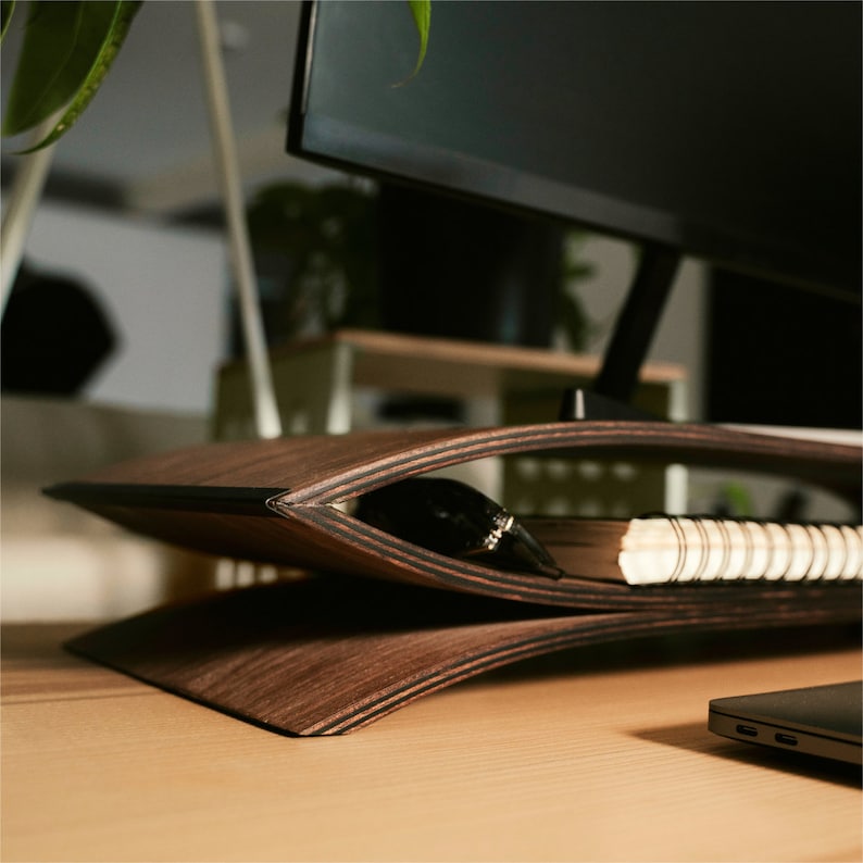 May include: A dark brown, curved wooden monitor stand with a layered plywood edge design. The stand features a hollow center space currently holding a spiral-bound notebook and a small black object, supporting a computer monitor above.