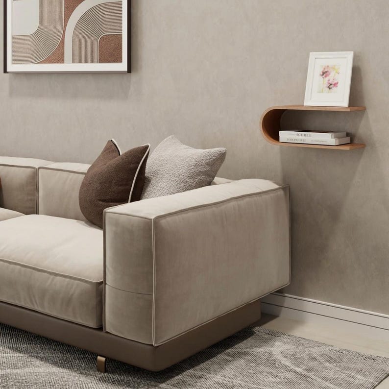 May include: A modern living room scene featuring a beige sectional sofa with brown accent pillows. A wooden floating shelf displays a framed floral print and books. A framed abstract art piece hangs on the wall.
