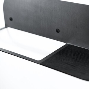 May include: A black metal shelf with a white rectangular shelf and a black wooden shelf. The shelf is mounted to a wall.
