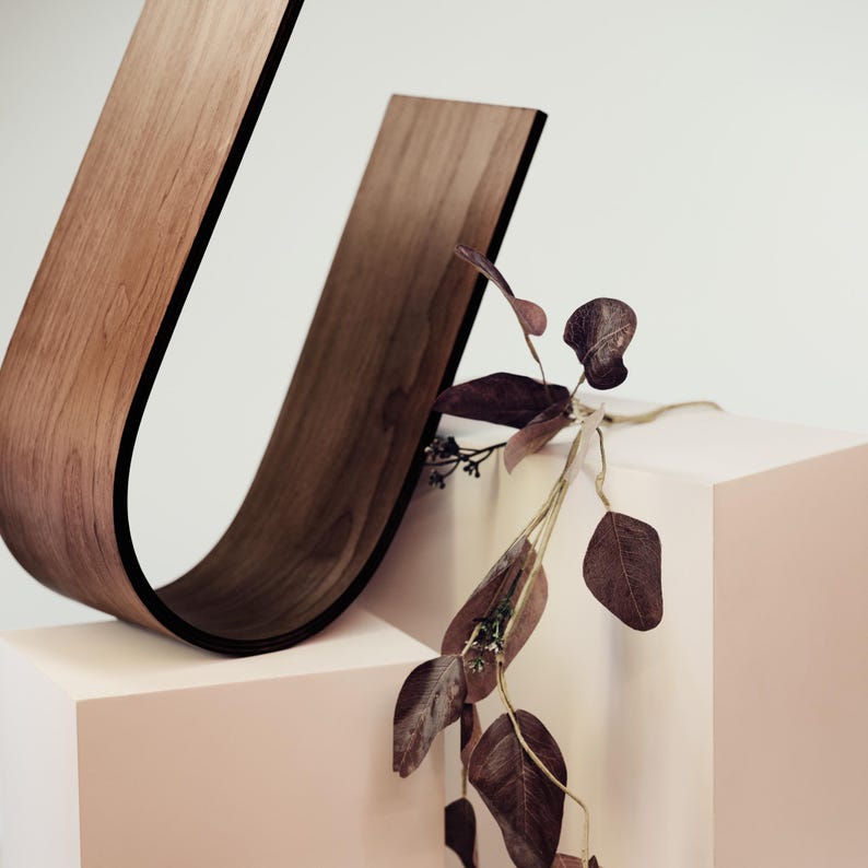 May include: A modern wooden sculpture in a U-shape with a dark edge, displayed on a light pink cube. Dried, burgundy-colored leaves and stems add a natural accent to the minimalist design. The sculpture's wood grain is visible.