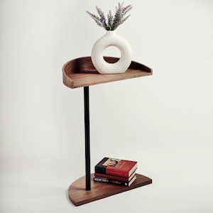 Puede incluir: Una mesa auxiliar moderna de dos niveles con acabado en madera oscura. La repisa superior sostiene un jarrón blanco circular con flores moradas. La repisa inferior exhibe dos libros. Un poste de metal negro sostiene las repisas.