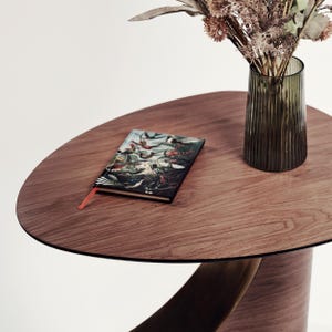 May include: A round wooden table with a dark brown finish. A small book with a colorful illustration and an orange bookmark sits on the table. A green glass vase with dried flowers is also on the table.