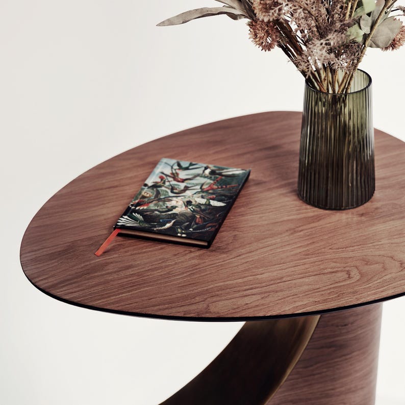 May include: A round wooden table with a dark brown finish. A small book with a colorful illustration and an orange bookmark sits on the table. A green glass vase with dried flowers is also on the table.