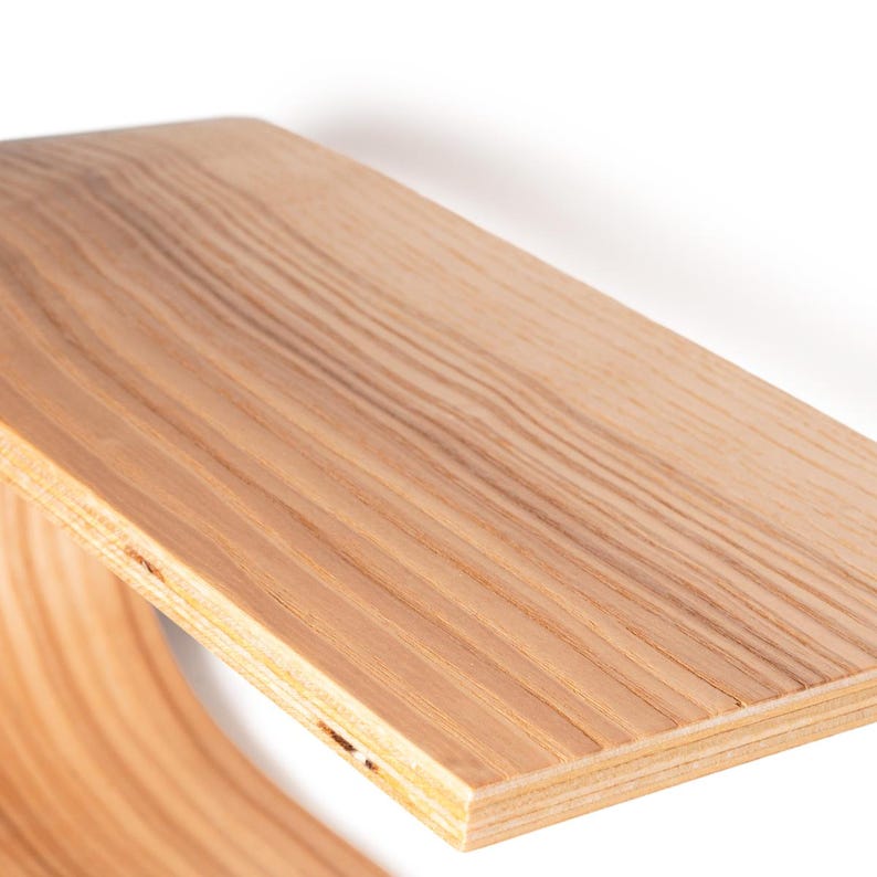 May include: Close-up of a wooden shelf with a curved support. The shelf is made of light-colored wood with visible grain patterns. The design features a smooth, curved base and a flat top, showcasing a minimalist aesthetic.