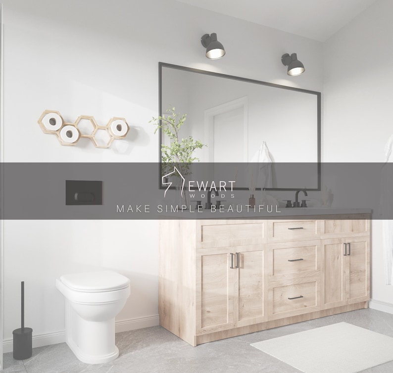 May include: A bathroom interior featuring a light wood vanity with black hardware, a large rectangular mirror, and two wall-mounted lights. A white toilet sits near a wall-mounted hexagonal wooden toilet paper holder. The text Ewart Woods Make Simple Beautiful is visible.