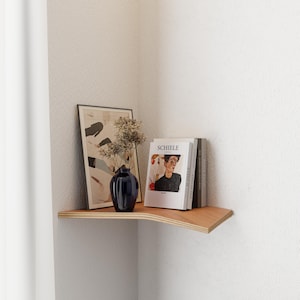 Op de afbeelding: Een houten hoekplank toont een ingelijste kunstdruk, een donkerblauwe vaas met gedroogde bloemen en verschillende boeken, waaronder een met de titel "SCHIELE". De plank is aan een witte muur bevestigd.