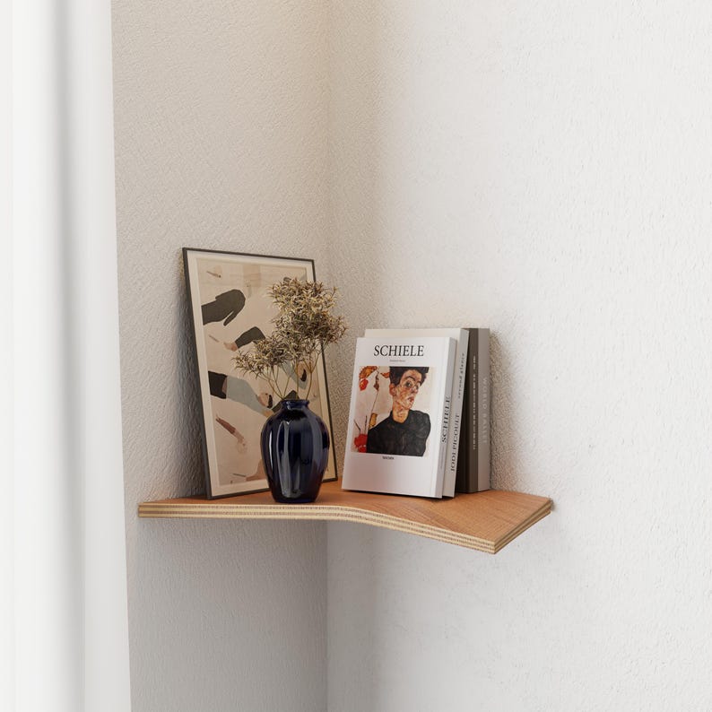 Op de afbeelding: Een houten hoekplank toont een ingelijste kunstdruk, een donkerblauwe vaas met gedroogde bloemen en verschillende boeken, waaronder een met de titel "SCHIELE". De plank is aan een witte muur bevestigd.
