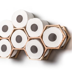 May include: A wall-mounted toilet paper holder constructed from light-colored wood with a honeycomb-inspired hexagonal frame. The structure features multiple interconnected compartments, each holding a single roll of white toilet paper.