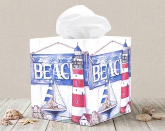 May include: A square tissue box with a nautical theme. The box features a lighthouse, sailboat, and the words "To the Beach" in blue and white. The lighthouse is red and white, and the sailboat has a blue and white sail. White tissues are visible at the top.