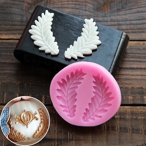 May include: Two silicone molds for baking, each with a design of two overlapping fern leaves. The molds are pink and are sitting on a wooden surface.