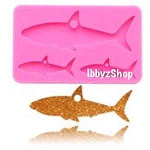 May include: Pink silicone mold with three shark shapes. The mold is for making resin or polymer clay crafts. The largest shark is about 3 inches long. The mold is labeled "IbbyzShop".