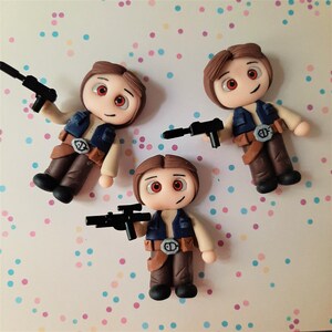 Può includere: Tre figurine fatte a mano in argilla polimerica di un personaggio con i capelli castani, che tiene in mano un blaster nero. Le figure indossano un gilet blu, una camicia beige, pantaloni marroni e una cintura con fibbia argentata. Lo sfondo è rosa chiaro con pois colorati.