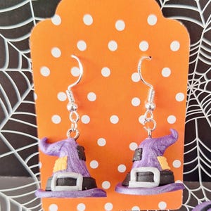 Puede incluir: Un par de pendientes de Halloween con pequeños sombreros de bruja morados con hebillas negras y detalles amarillos. Los pendientes cuelgan de alambres plateados en forma de anzuelo. Los pendientes se exhiben en una tarjeta naranja con lunares blancos, sobre un fondo de telaraña.