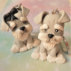 Puede incluir: Dos llaveros hechos a mano con perros Schnauzer. Los perros son grises y blancos con narices y ojos negros. Un llavero tiene un lazo negro. Sobre un fondo pastel.
