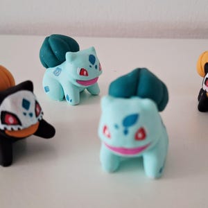 Op de afbeelding: Vier kleine beeldjes van een populair personage. Twee zijn lichtblauw met groene toppen en twee zijn zwart met oranje pompoenachtige toppen. De beeldjes hebben rode, witte en blauwe accenten.