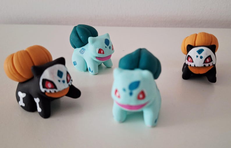 Op de afbeelding: Vier kleine beeldjes van een populair personage. Twee zijn lichtblauw met groene toppen en twee zijn zwart met oranje pompoenachtige toppen. De beeldjes hebben rode, witte en blauwe accenten.