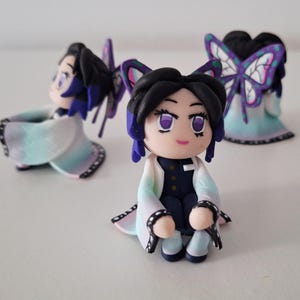 May include: Handmade polymer clay figurines of a character with black hair, purple eyes, and butterfly hair accessories. The figurines wear a white and blue kimono with black accents. The figures are posed in a sitting position.