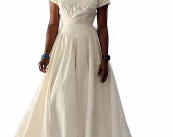 May include: Ivory-colored wedding dress with a fitted bodice and a full skirt. The dress features cap sleeves and floral embellishments along the neckline. The skirt flows to the floor, creating an elegant silhouette.