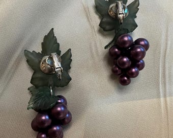 May include: A pair of dangle earrings featuring a cluster of purple grapes hanging from a green leaf design. The earrings are made of plastic and have a silver-toned clip-on closure.