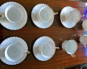 May include: A set of ten white teacups and saucers with gold trim. The cups and saucers are arranged in two rows of five on a wooden surface.