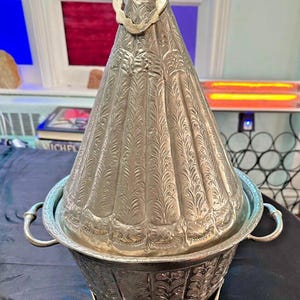 May include: A silver serving dish with a lid and a decorative handle. The dish has an ornate design and sits on three feet. The lid is shaped like a cone and has a decorative knob on top.