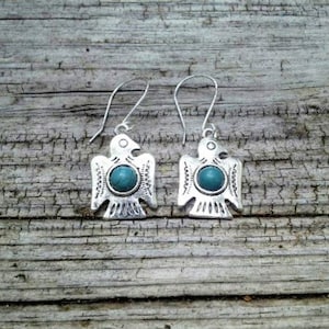 May include: A pair of silver-toned earrings featuring a stylized bird design with a turquoise-colored stone in the center. The earrings are suspended from a simple hook. The background is a weathered wooden surface.