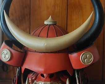May include: A red and gold samurai helmet with horns. The helmet has a fierce, stylized face mask with large teeth. The horns are black and gold, and the helmet is decorated with a gold symbol.