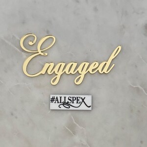 May include: A gold-coloured 'Engaged' word cutout in a cursive font, with a small rectangular sign below that reads '#ALLSPEX' in black text on a white background. The background is a light grey marble.