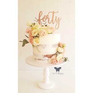 May include: A two-tiered cake with a semi-naked frosting design, decorated with fresh pink and cream roses and greenery. A rose gold "forty" cake topper sits on top. The cake is presented on a white cake stand.