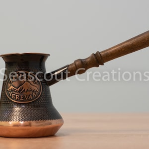 May include: A traditional copper coffee pot with a wooden handle. The pot has a black and copper finish and features an embossed design with the words "Armenia Yerevan".