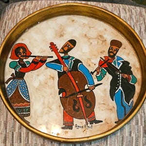 May include: Round serving tray with a gold rim and a painted scene of three musicians. The musicians are playing a flute, cello, and violin. The tray has a cream-colored background with a marbled effect. The artist's signature, "Amos Amit," is visible.