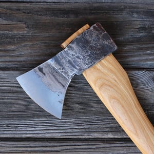 Könnte beinhalten: Eine Axt mit einer verwitterten Stahlklinge und einem hellen Holzgriff. Der Axtkopf ist mit einem Holzkeil am Griff befestigt. Die Axt liegt auf einem Hintergrund aus gealterten, grauen Holzplanken.