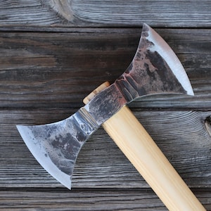 May include: A double-bladed axe with a textured metal head and a light wooden handle. The axe head has a dark, mottled appearance with a sharp edge. The handle is made of light-coloured wood. It rests on a weathered wooden surface.