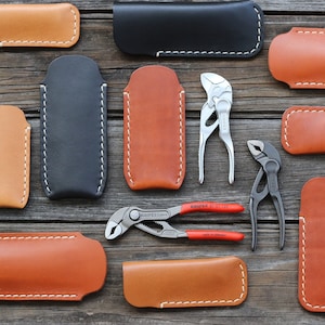 Può includere: Una collezione di 12 portautensili in pelle in varie tonalità di marrone e nero, con cuciture bianche. I portautensili sono disposti su una superficie di legno. Nell'immagine sono inclusi anche due paia di pinze.