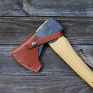 Könnte beinhalten: Eine braune Lederaxt-Scheide mit einem Metall-Druckknopfverschluss und einer Axt mit Holzstiel, auf dem der Markenname "Hultafors" aufgedruckt ist.
