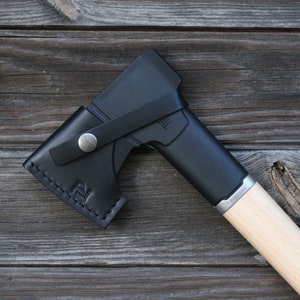 May include: A black axe with a wooden handle and a leather sheath. The axe has a silver metal band around the handle with the word "Fiskars" printed on it.