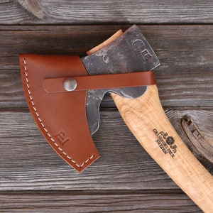 Könnte beinhalten: Eine braune Lederscheide mit einem Metall-Druckknopfverschluss und einem kleinen Metall-Logo auf der Vorderseite. Die Scheide hält eine kleine Axt mit einem Holzstiel. Die Axt hat einen Metallkopf mit einer gebogenen Klinge und einem kleinen Metall-Logo an der Seite. Die Axt wird von Gränsfors Bruk in Schweden hergestellt.