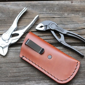 Könnte beinhalten: Zwei Metallzangen und eine braune Ledertasche mit schwarzem Clip. Eine Zange ist silberfarben, die andere dunkelgrau. Die Zangen sind mit "KNIPEX" beschriftet. Die Ledertasche hat einen Knopfverschluss und genähte Kanten.