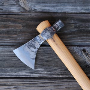 May include: A handcrafted axe with a wooden handle and a silver-coloured blade. The axe head is attached to the handle with a metal piece. The axe is set against a weathered wooden background.