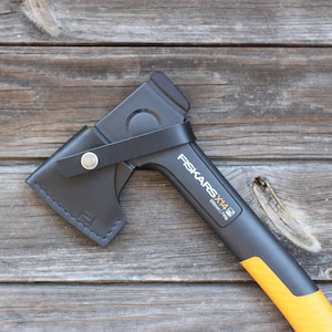 Może przedstawiać: Czarna siekiera Fiskars X14 z czarną skórzaną pochwą. Siekiera ma żółty trzonek, a na nim nadrukowany jest napis "Fiskars X14".