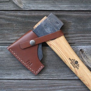 May include: A handcrafted axe with a wooden handle and a metal blade, protected by a brown leather sheath. The axe head is marked with the initials "HF". The handle is engraved with "Gransfors Bruk Sweden".
