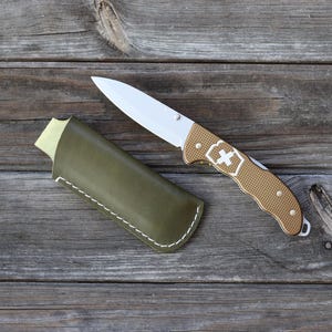 May include: A brown leather knife sheath with a white stitch detail and a silver and gold pocket knife with a white cross logo.