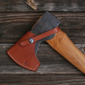 Könnte beinhalten: Eine braune Lederaxt-Scheide mit einem Metallknopf und einem gestickten Design. Die Scheide ist an einem Axt-Stiel aus Holz befestigt.