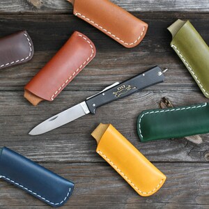 Könnte beinhalten: Eine Sammlung von Leder-Messerhüllen in verschiedenen Farben, darunter Braun, Orange, Grün, Blau und Gelb. Die Hüllen haben weiße Nähte und werden auf einer Holzoberfläche präsentiert. Ein kleines schwarzes Klappmesser mit einem Holzgriff ist ebenfalls abgebildet.