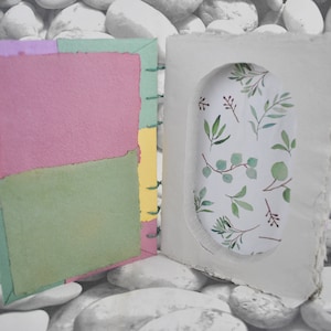 May include: Handmade journal with a textured cover and colorful pages. The open journal reveals a pink and green page, with a decorative oval cutout showcasing a botanical print of green leaves and branches on a white background. The journal is bound with green thread.