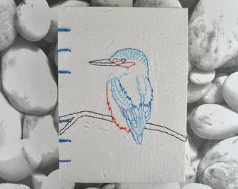 May include: A small, square, white canvas with a hand-stitched illustration of a kingfisher bird perched on a branch. The bird is outlined in blue and red thread. The canvas has blue stitching along the left edge.