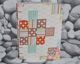 May include: A handmade journal with a patchwork design on the cover. The cover features squares of fabric in various patterns and colors, including red, brown, teal, and pink. The binding is stitched with red thread.