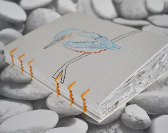 May include: A handmade journal with a bird design embroidered on the cover. The bird is blue and orange, with black stitching for the branch. The pages are held together with orange thread.